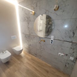 Ter-Bud - Nowoczesna łazienka z marmurowymi płytkami, okrągłym lustrem z złotymi akcentami, podwieszaną toaletą i bidetem, oświetleniem LED i drewnopodobną podłogą.