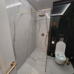 Ter-Bud - Elegancka łazienka z kabiną prysznicową typu walk-in z elementami w kolorze złotym, marmurowe płytki na ścianach i podłodze, nowoczesna toaleta myjąca z pozłacanym koszem na śmieci i czarnym...
