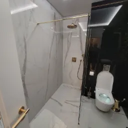 Elegancka łazienka z kabiną prysznicową typu walk-in z elementami w kolorze złotym, marmurowe płytki na ścianach i podłodze, nowoczesna toaleta myjąca z pozłacanym koszem na śmieci i czarnym...