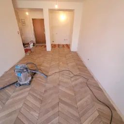 Nowo ułożona podłoga z drewnianych paneli w jodełkę, odkurzacz przemysłowy w rogu, przygotowania do dalszych prac wykończeniowych w tle.