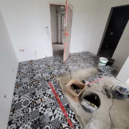 Układanie czarno-białych płytek podłogowych w geometryczne wzory, widoczne narzędzia glazurnicze i poziomica, fragmenty podłogi z fugą, prace w toku w nowym mieszkaniu.