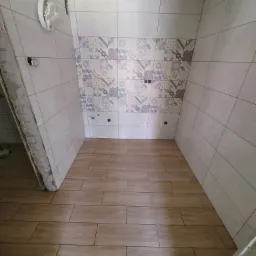 Wnętrze łazienki w trakcie remontu: położone płytki podłogowe imitujące drewno, częściowo wykończone płytki ścienne z dekoracyjnym pasem, widoczne otwory na instalacje.