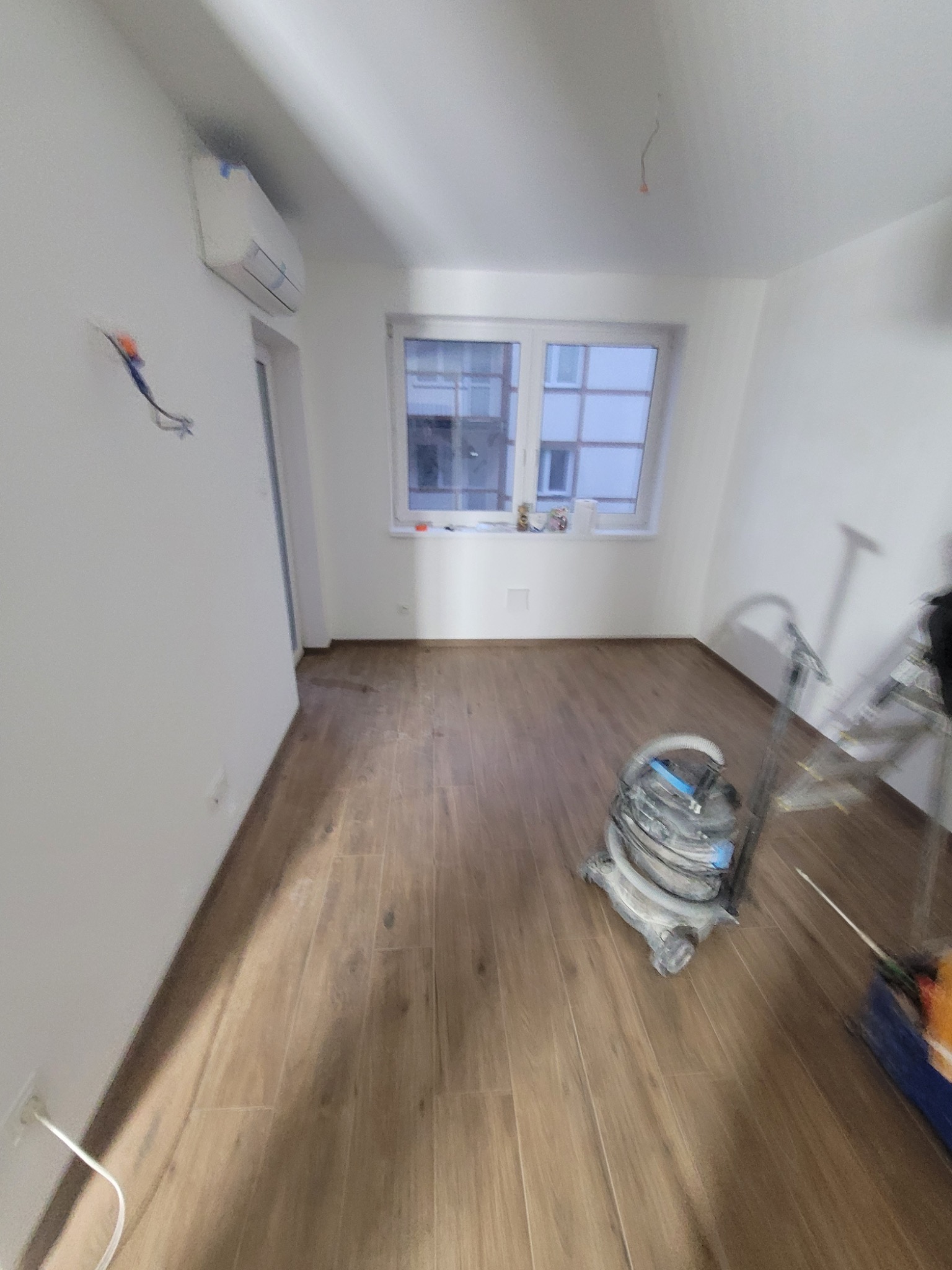 Pokój po ułożeniu paneli podłogowych z widocznymi niedokończonymi pracami elektrycznymi, klimatyzacją, oknem i sprzętem do sprzątania.