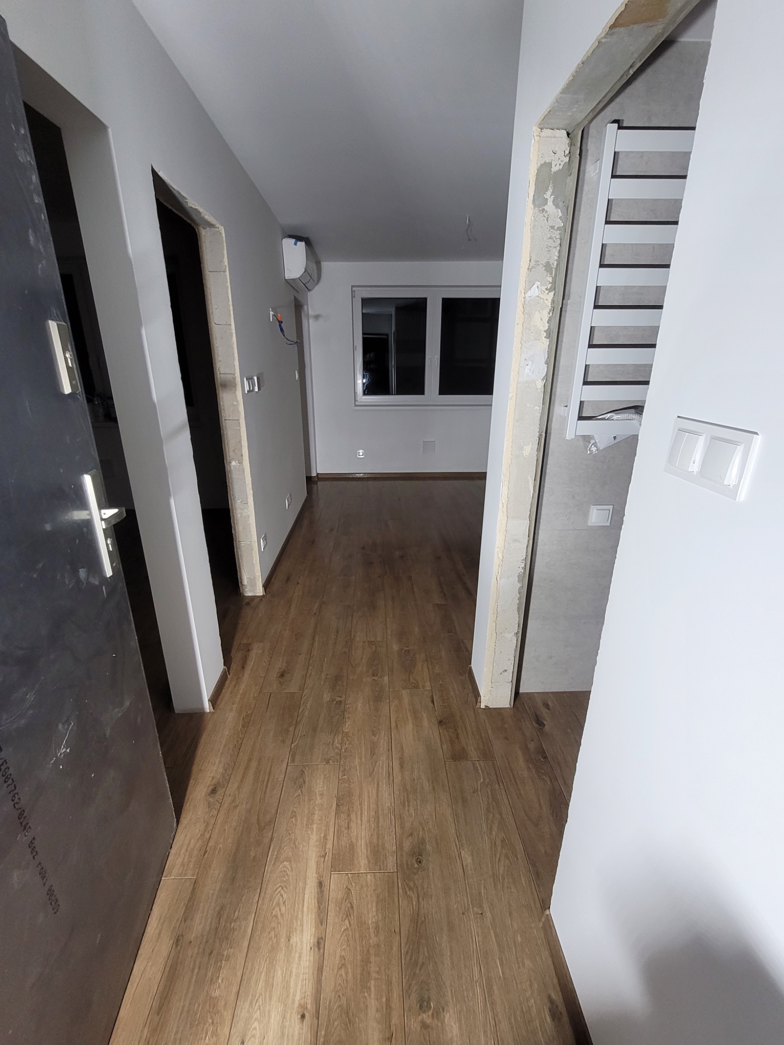 Wnętrze mieszkania w trakcie prac wykończeniowych: widoczne panele podłogowe, ściany przygotowane do malowania, surowa ościeżnica i grzejnik łazienkowy w tle.