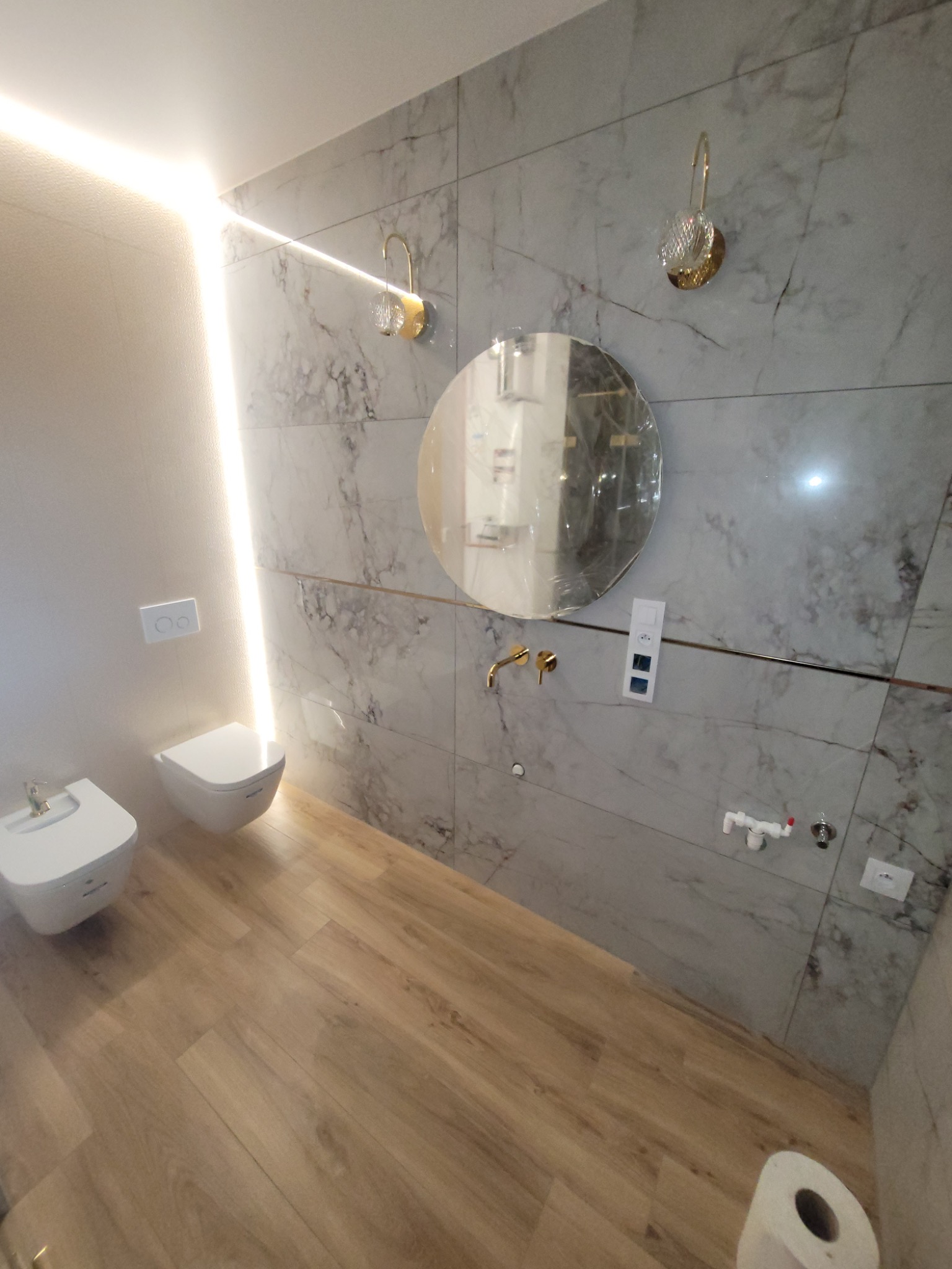 Nowoczesna łazienka z marmurowymi płytkami, okrągłym lustrem z złotymi akcentami, podwieszaną toaletą i bidetem, oświetleniem LED i drewnopodobną podłogą.