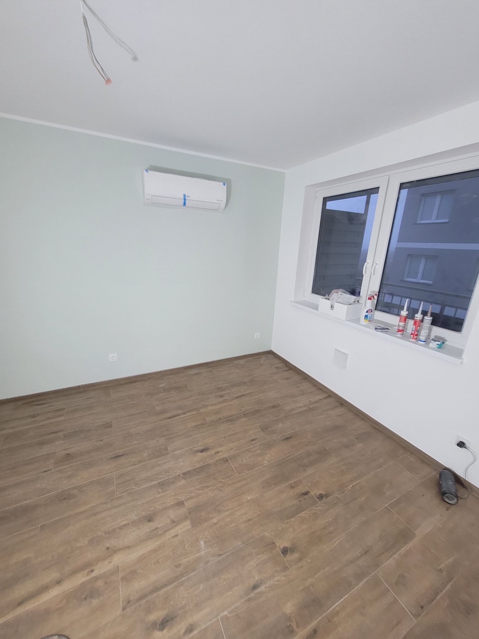 Pokój po odświeżeniu: świeżo położone panele podłogowe imitujące drewno, pomalowane na jasnozielono ściany z zamontowanym klimatyzatorem, białe okno z widokiem na blok.