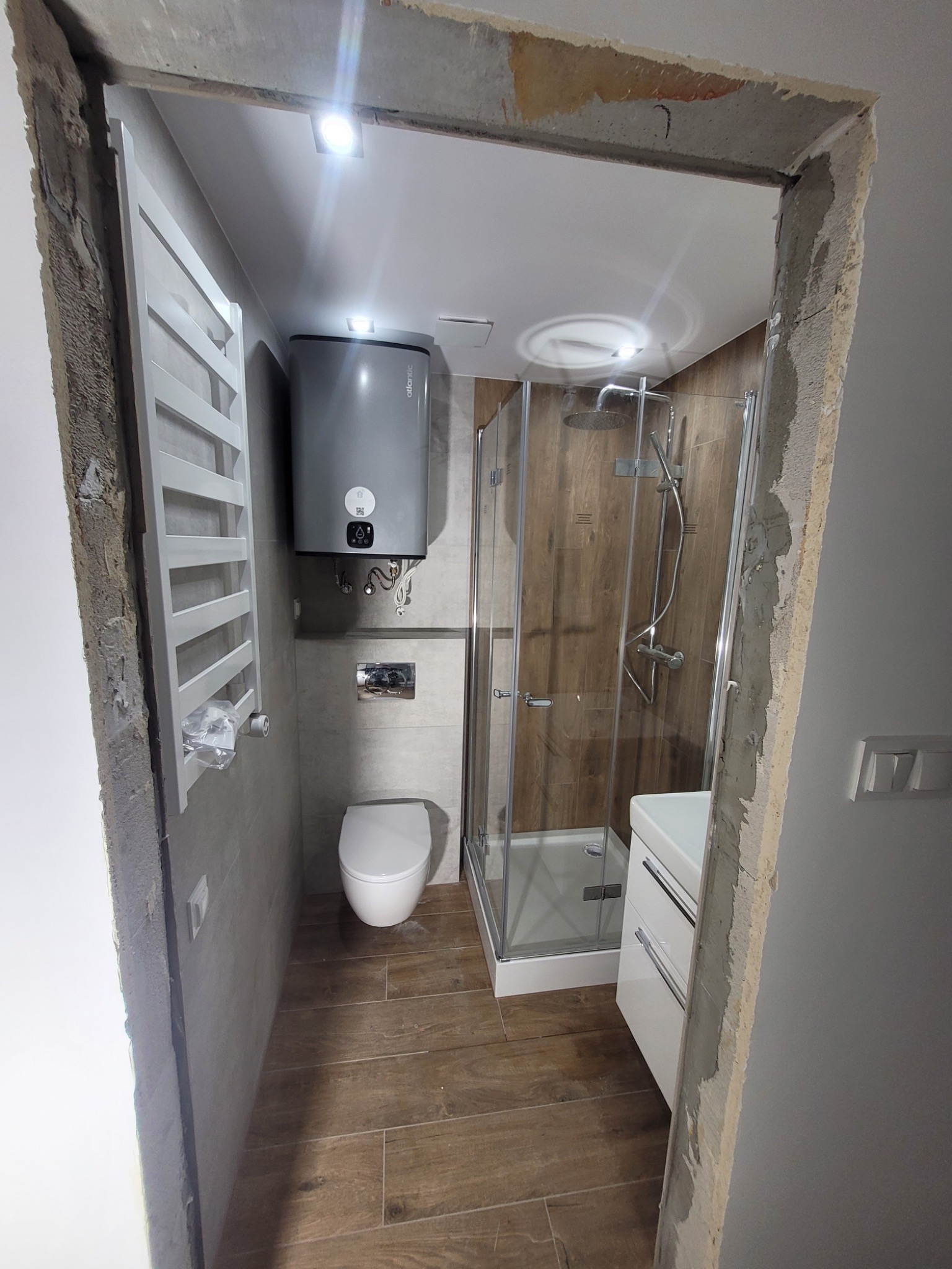 Nowoczesna łazienka z kabiną prysznicową, grzejnikiem drabinkowym i podgrzewaczem wody, widoczna przez surowe, nieotynkowane obramowanie drzwi.