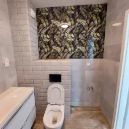 Wykończona łazienka z wiszącą toaletą, białą szafką z umywalką i ścianą z płytek z motywem liści bananowca.