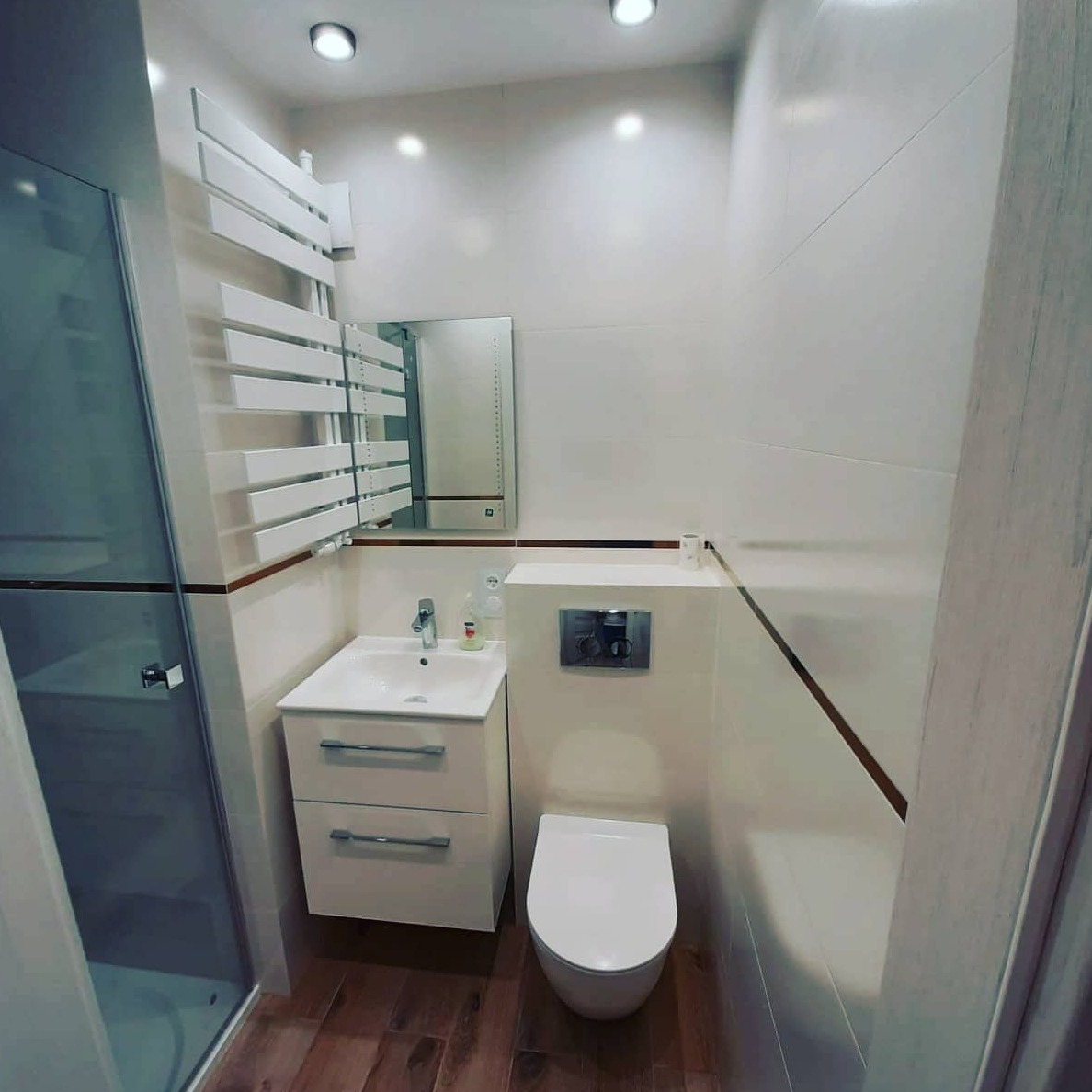 Nowoczesna, jasna łazienka po remoncie: białe płytki, umywalka z szafką, toaleta podwieszana z chromowanym przyciskiem, grzejnik drabinkowy i kabina prysznicowa ze szkła.