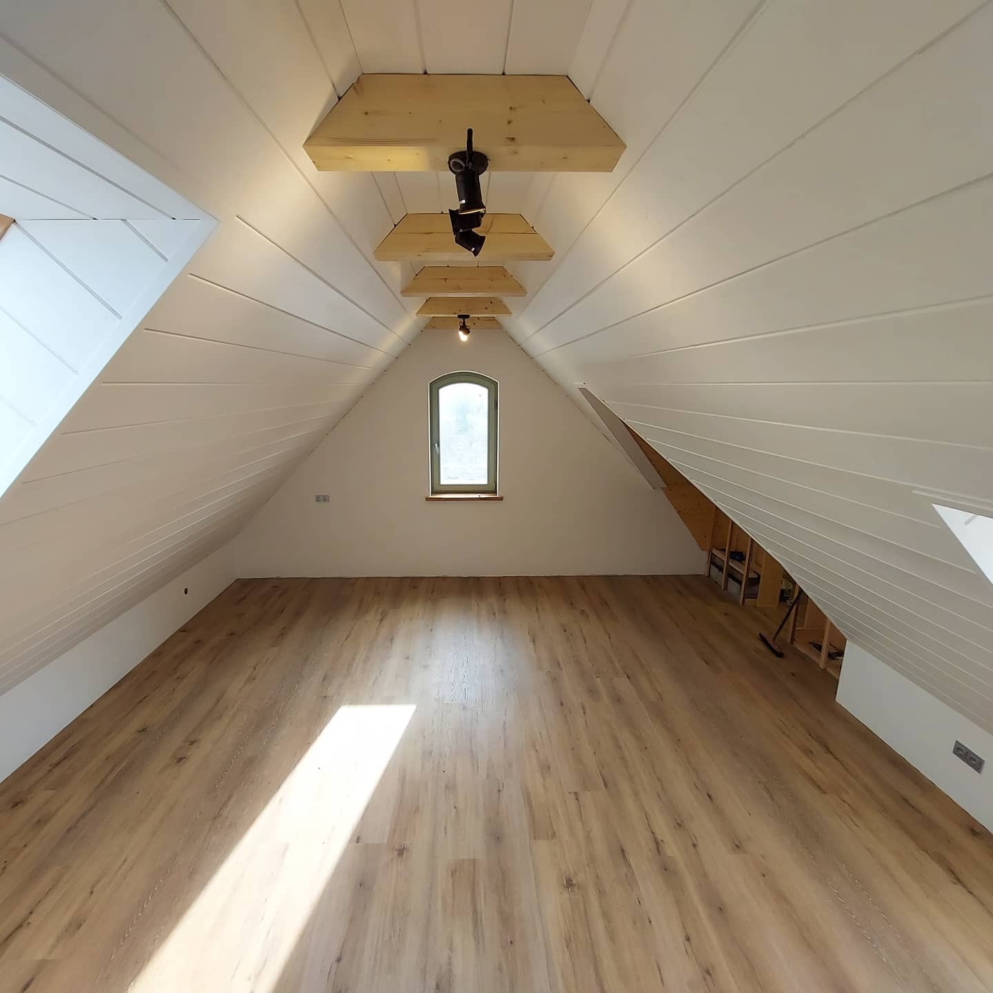 Jasne, puste poddasze po remoncie z drewnianą podłogą i białymi panelami na skośnych ścianach, oświetlone reflektorami zawieszonymi na drewnianych belkach.