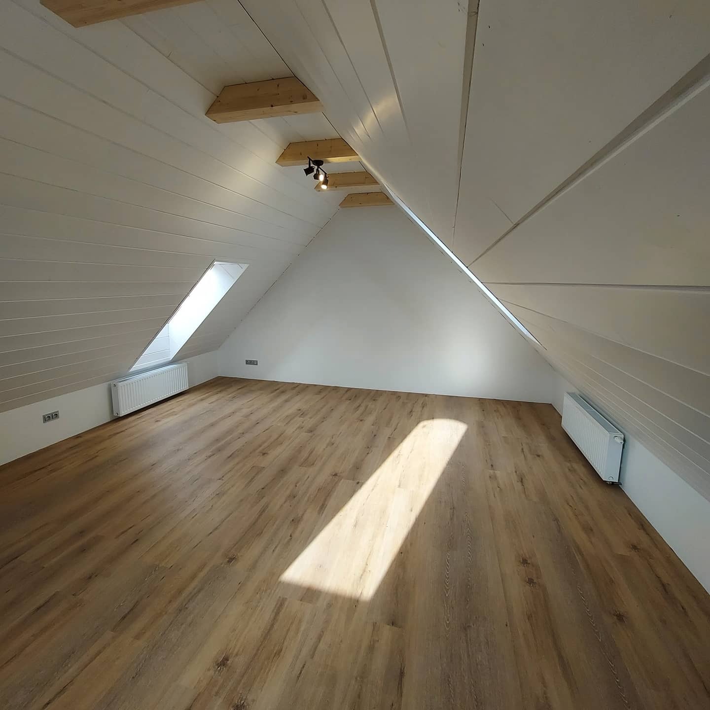 Poddasze po remoncie z nową podłogą imitującą drewno, skośnym oknem w dachu i białymi grzejnikami pod oknem.