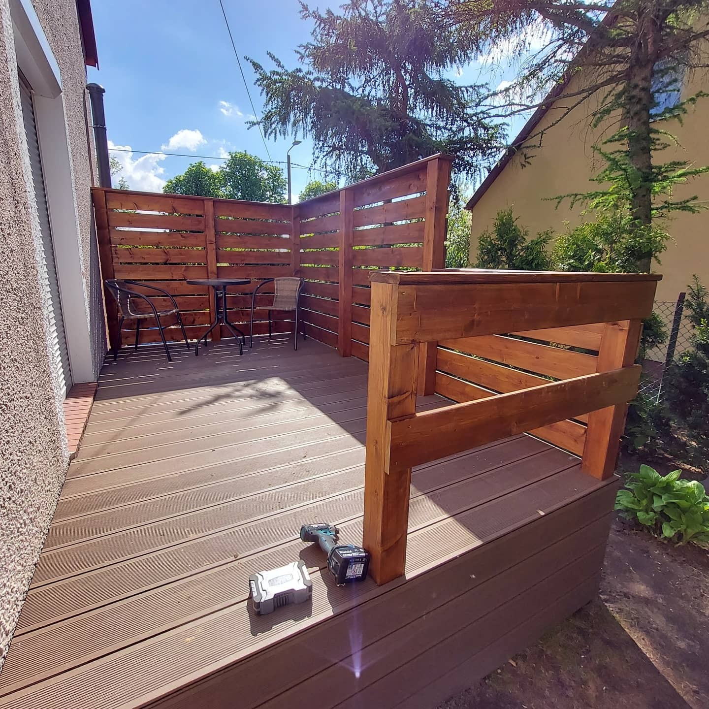 Nowoczesny taras z drewnianą podłogą i ażurową zabudową, widoczne narzędzia akumulatorowe, stół i krzesła na tle zieleni.