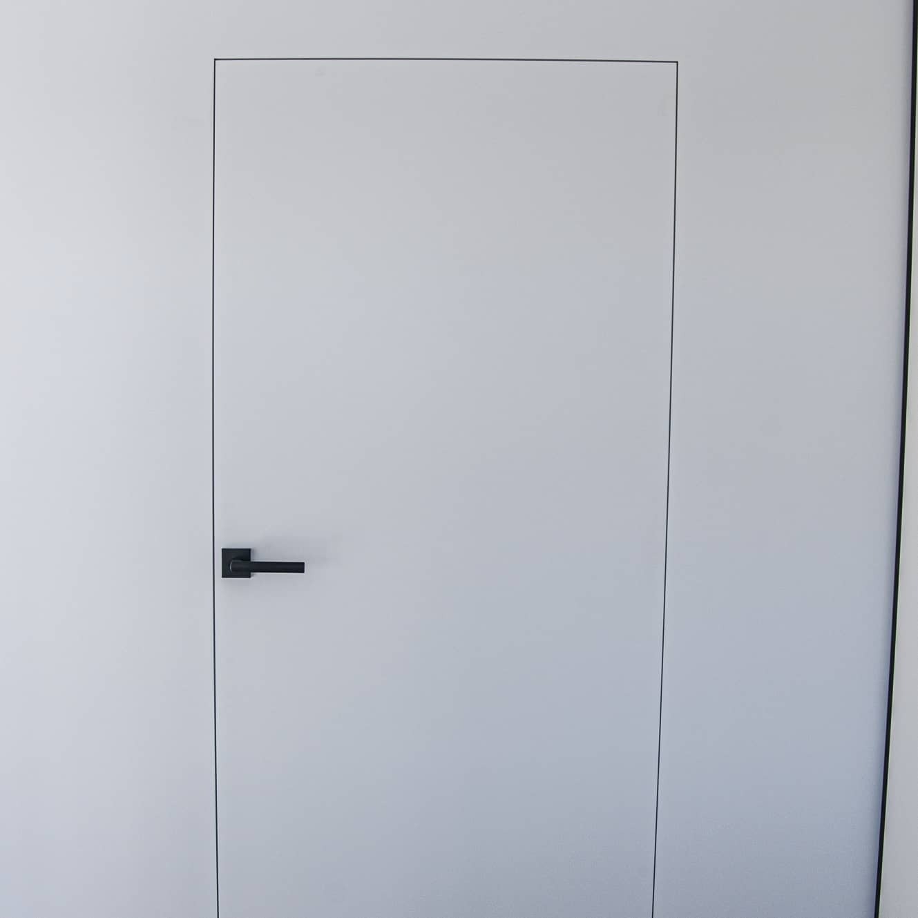 Minimalistyczne białe drzwi wewnętrzne z ukrytą ościeżnicą i czarną, kwadratową klamką, wkomponowane w białą ścianę.