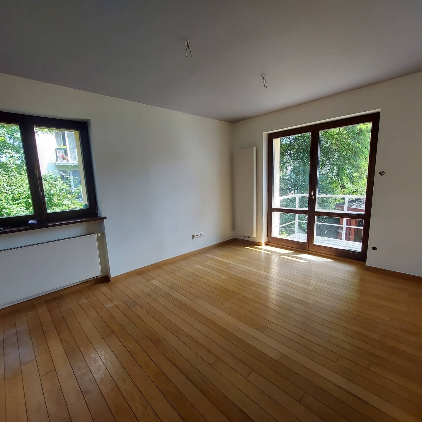 Pusty pokój z drewnianą podłogą, białymi ścianami i dużymi oknami z widokiem na zieleń. Widoczne gniazdka elektryczne i wystający kabel ze ściany.