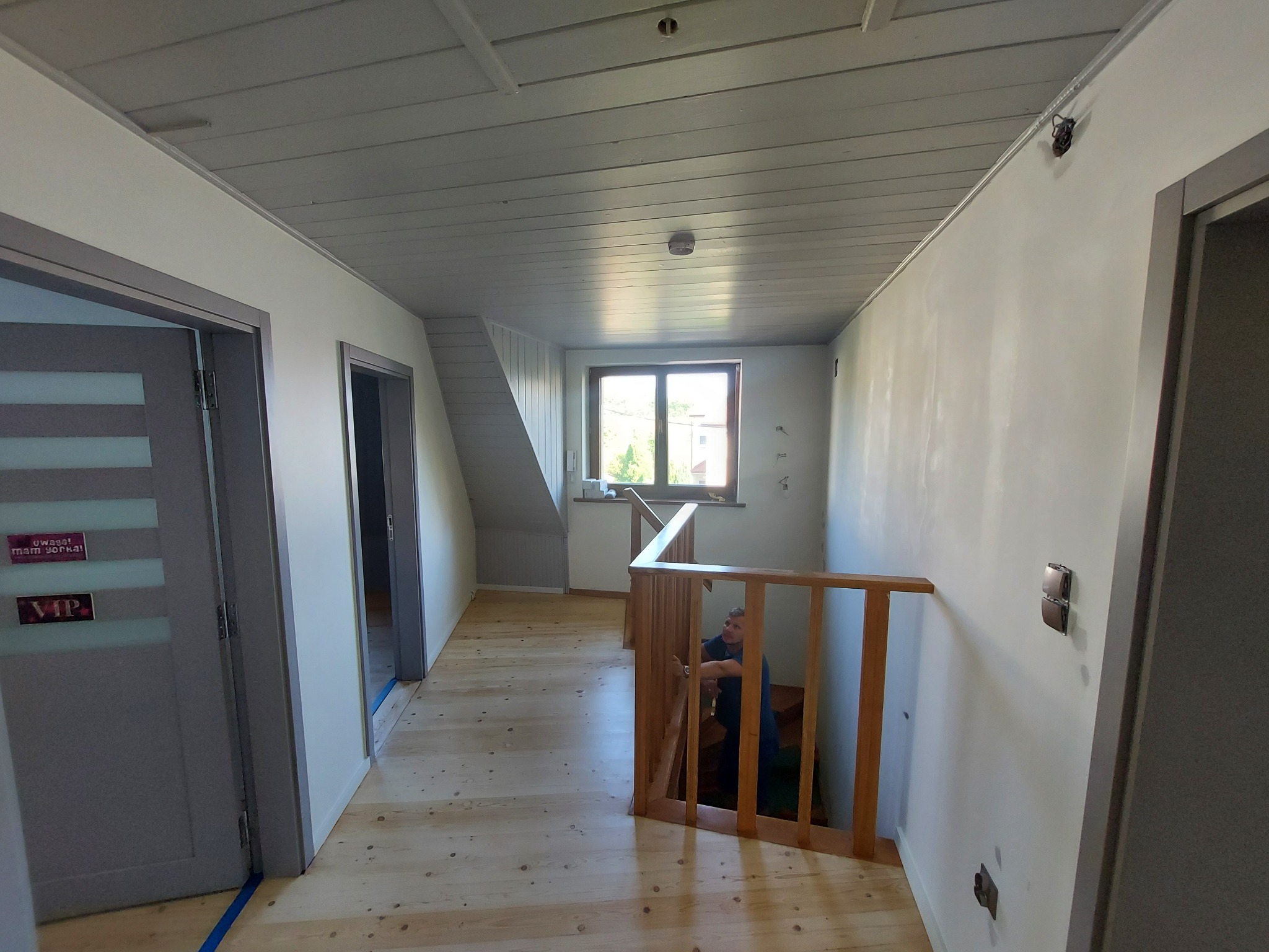 Korytarz w domu jednorodzinnym z drewnianą podłogą, szarym sufitem i białymi ścianami; widoczny stolarz montujący drewniane schody, częściowo zasłonięty przez balustradę.
