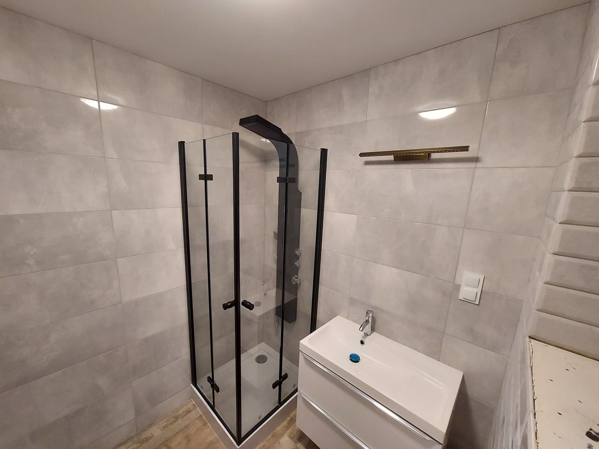 Nowoczesna łazienka z kabiną prysznicową typu walk-in z czarnymi profilami, białą umywalką z szafką i szarymi płytkami imitującymi beton.