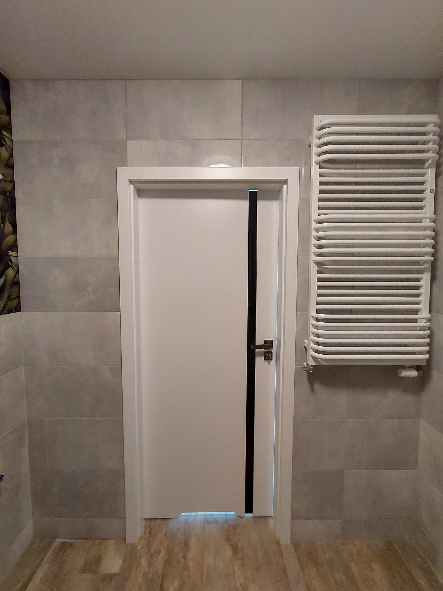 Nowoczesna łazienka z białymi płytkami imitującymi kamień, białą drzwiami z czarnym akcentem i grzejnikiem drabinkowym.