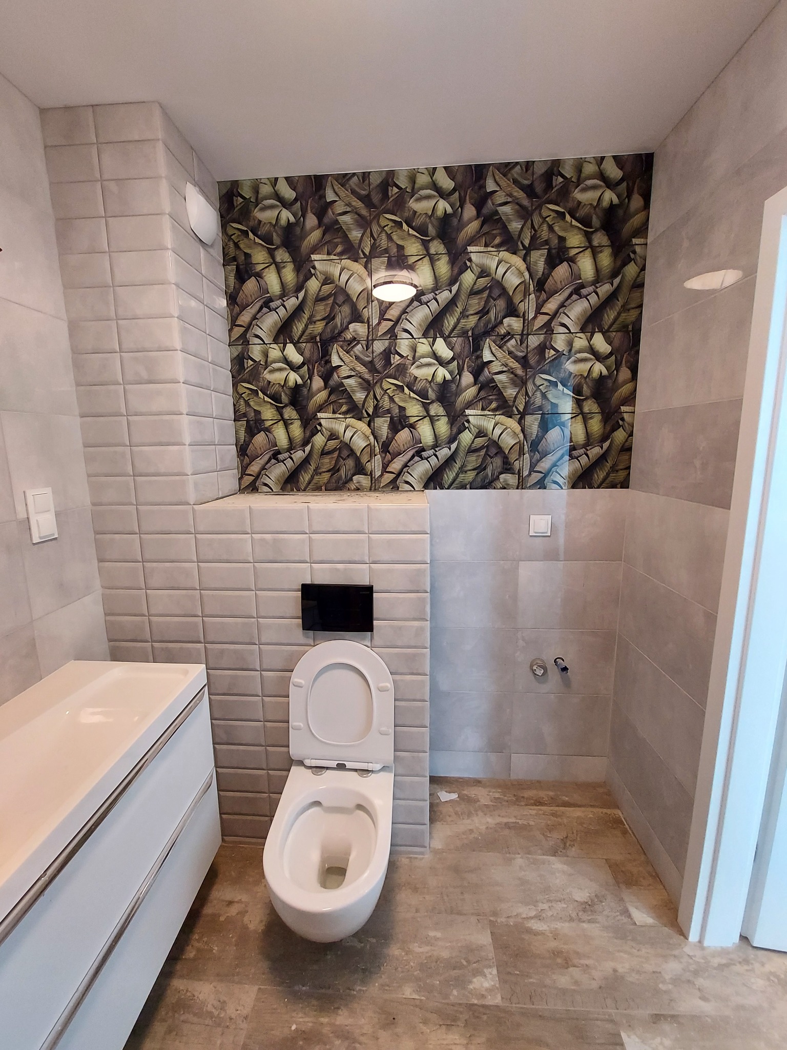 Wykończona łazienka z wiszącą toaletą, białą szafką z umywalką i ścianą z płytek z motywem liści bananowca.