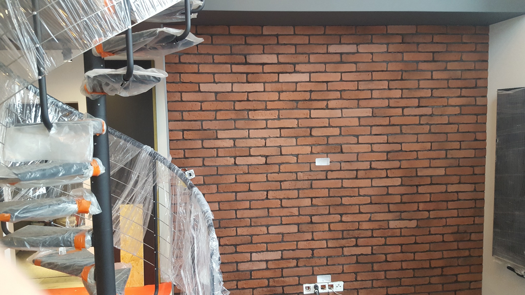 Ceglana ściana dekoracyjna z widoczną instalacją elektryczną i spiralnymi schodami zabezpieczonymi folią malarską.