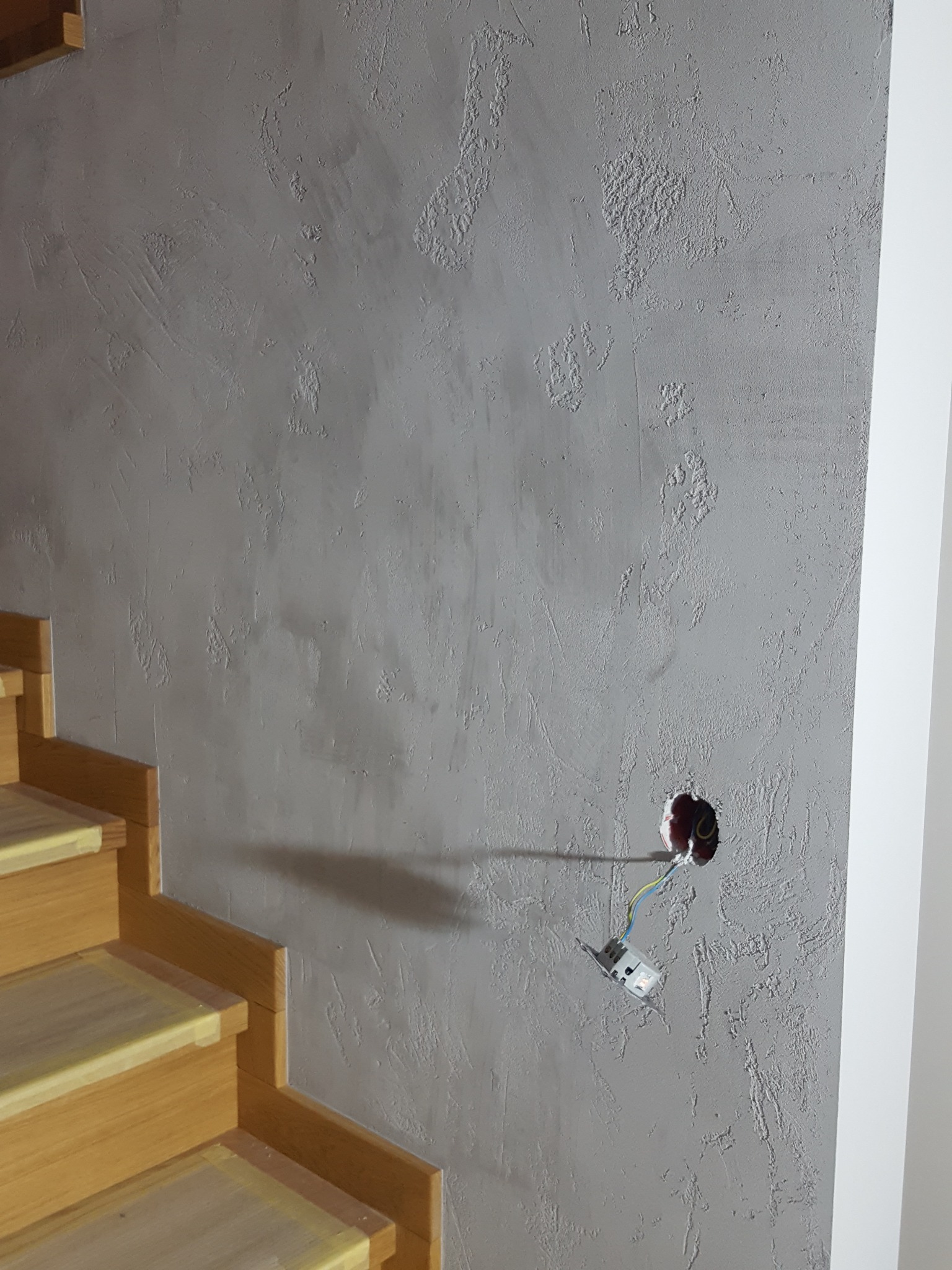 Szara ściana z dekoracyjnym tynkiem strukturalnym w trakcie remontu, widoczne drewniane schody i wystająca puszka elektryczna z przewodami.