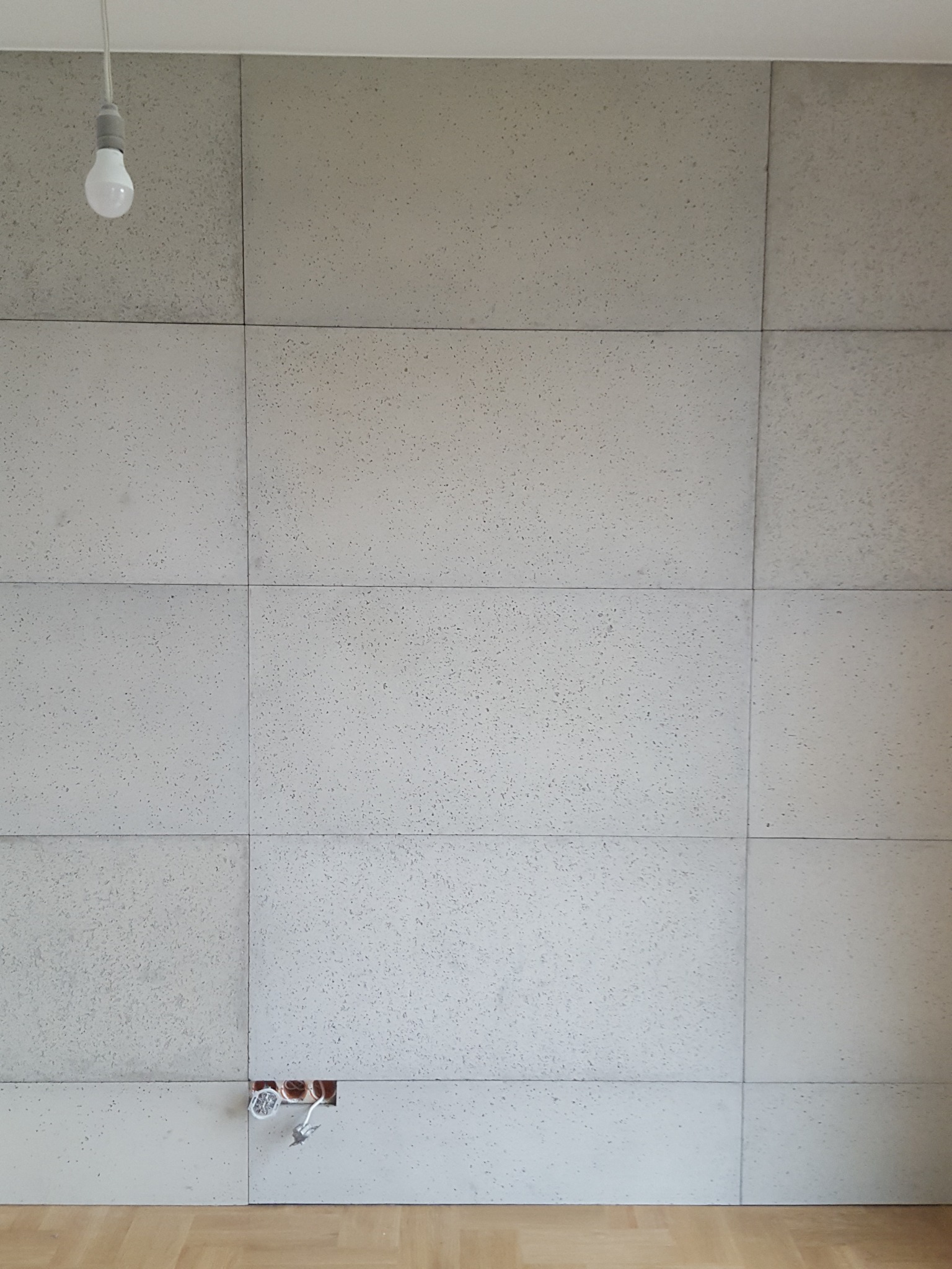 Ściana wykończona panelami betonowymi z widocznymi kablami elektrycznymi w otworze, oświetlona wiszącą żarówką.