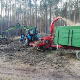 Niebieski traktor z zielonym ramieniem chwytającym gałęzie w lesie, obok czerwonej rębarki do drewna, która wyrzuca zrębki do zielonej przyczepy.