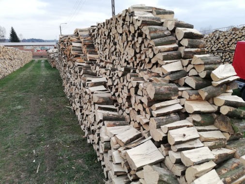 Rząd ułożonego drewna opałowego na trawiastym terenie, z widocznym drugim rzędem w oddali i fragmentem czerwonego pojazdu po prawej stronie.