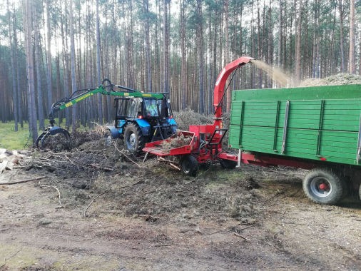 Niebieski traktor z zielonym ramieniem chwytającym gałęzie w lesie, obok czerwonej rębarki do drewna, która wyrzuca zrębki do zielonej przyczepy.