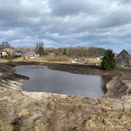 Nowo wykopany staw na tle wiejskiego krajobrazu z pochmurnym niebem, widoczne ślady po pracy maszyn ziemnych.
