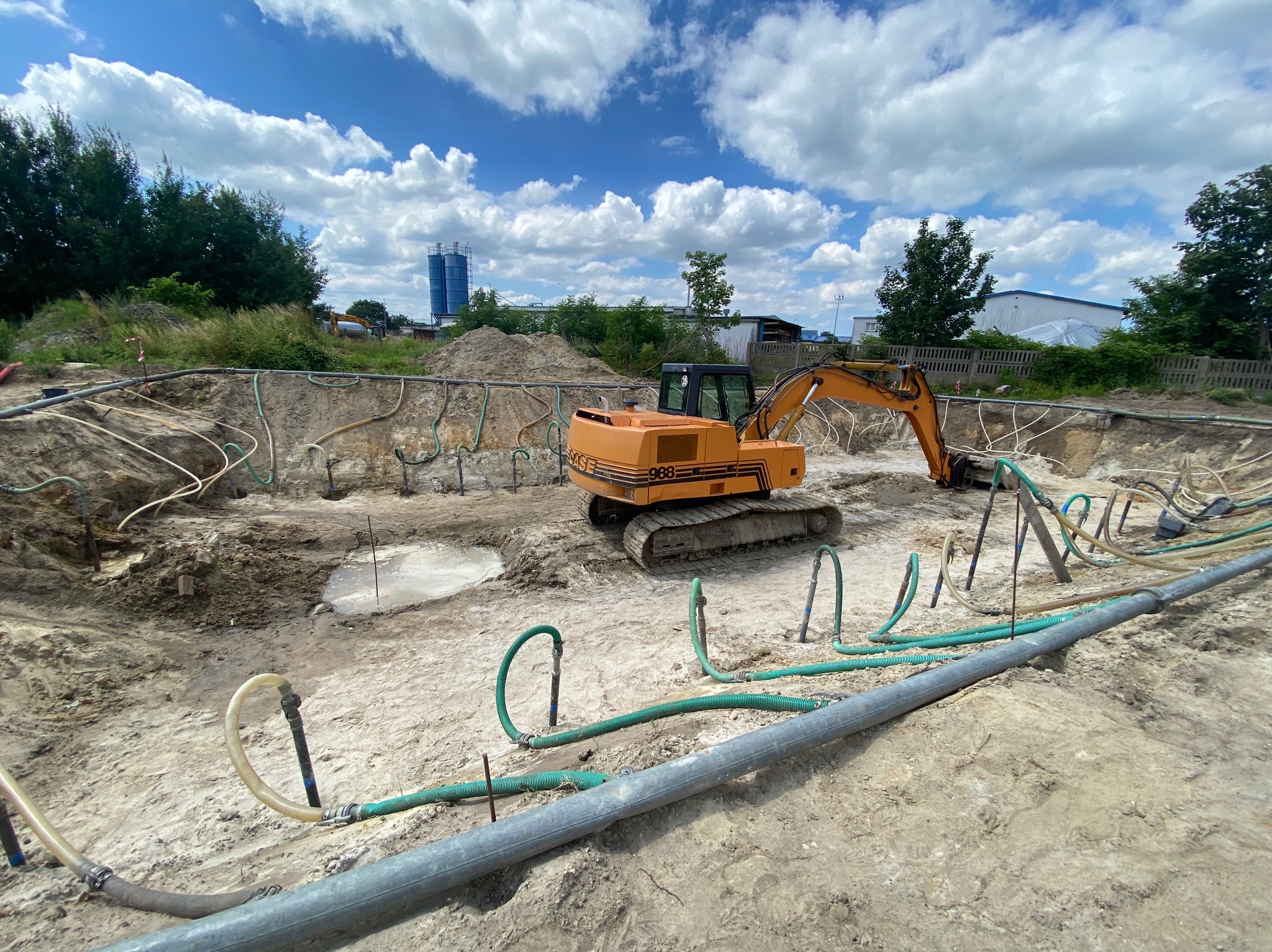 Pomarańczowa koparka gąsienicowa w wykopie, liczne węże i rury do odwadniania gruntu, błękitne niebo z chmurami w tle.