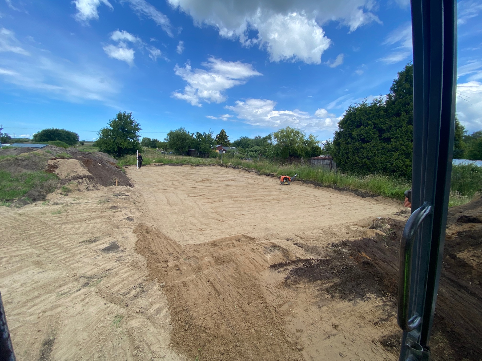 Przygotowany, wyrównany i ubity teren pod budowę, widok z kabiny maszyny, w tle błękitne niebo z chmurami i zieleń drzew.