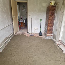 Kris Remonty wykończenia Złota Rączka - Wyrównana warstwa piasku na podłodze w pomieszczeniu w trakcie prac przygotowawczych, widoczny piec kaflowy, szpachelka, łopata i otwarta futryna drzwiowa.