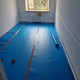 Przygotowanie do układania paneli: niebieska pianka podkładowa z pomarańczową taśmą, zwinięta rolka pianki i rolka taśmy klejącej w pokoju z grzejnikiem pod oknem.