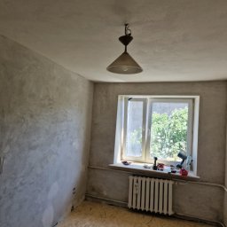 Kris Remonty wykończenia Złota Rączka - Pokój w trakcie odświeżania: ściany z surowym tynkiem, podłoga wyłożona płytami OSB, okno z widokiem na zieleń, grzejnik i narzędzia na parapecie, wisząca lampa.