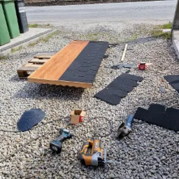 Przygotowanie drewnianej konstrukcji z bitumicznym pokryciem dachowym na palecie, narzędzia pracy leżą na żwirowym podłożu, w tle widoczne pojemniki na śmieci oraz pole rzepaku.