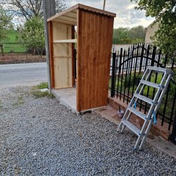 Kris Remonty wykończenia Złota Rączka - Drewniana konstrukcja z półkami, prawdopodobnie altana lub domek narzędziowy, w trakcie budowy, z drabinką opartą o ogrodzenie i wiaderkiem po farbie na żwirowym podłożu.