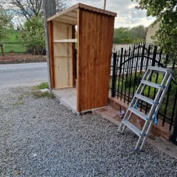 Drewniana konstrukcja z półkami, prawdopodobnie altana lub domek narzędziowy, w trakcie budowy, z drabinką opartą o ogrodzenie i wiaderkiem po farbie na żwirowym podłożu.