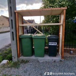 Kris Remonty wykończenia Złota Rączka - Zadaszona wiata na trzy pojemniki na śmieci: dwa zielone i jeden czarny z napisem 'Popiół', na żwirowym podłożu, z widocznym słupem energetycznym i fragmentem ogrodzenia.