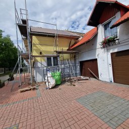 Kris Remonty wykończenia Złota Rączka - Dom jednorodzinny w trakcie prac remontowych elewacji i dachu, widoczne rusztowania, worki z materiałami budowlanymi i częściowo odnowiona fasada.