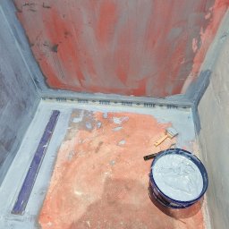 Kris Remonty wykończenia Złota Rączka - Przygotowanie podłoża w kabinie prysznicowej: czerwona hydroizolacja z widocznymi pociągnięciami pędzla na ścianach i podłodze, taśma uszczelniająca ATLAS na łączeniach, wiadro z szarą masą...