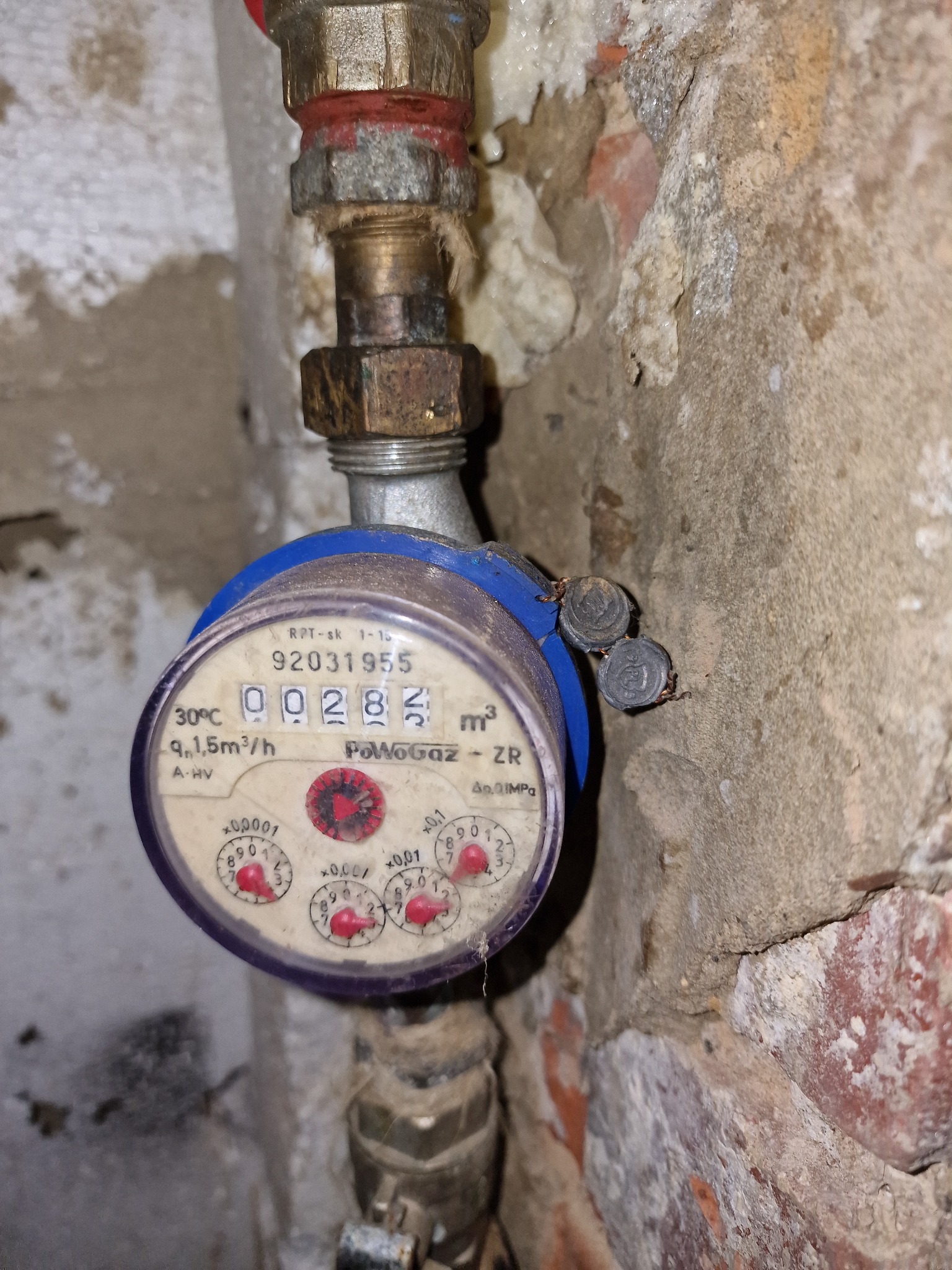 Licznik gazu z plombami na tle ściany z fragmentami cegły, widoczne złącza instalacyjne.