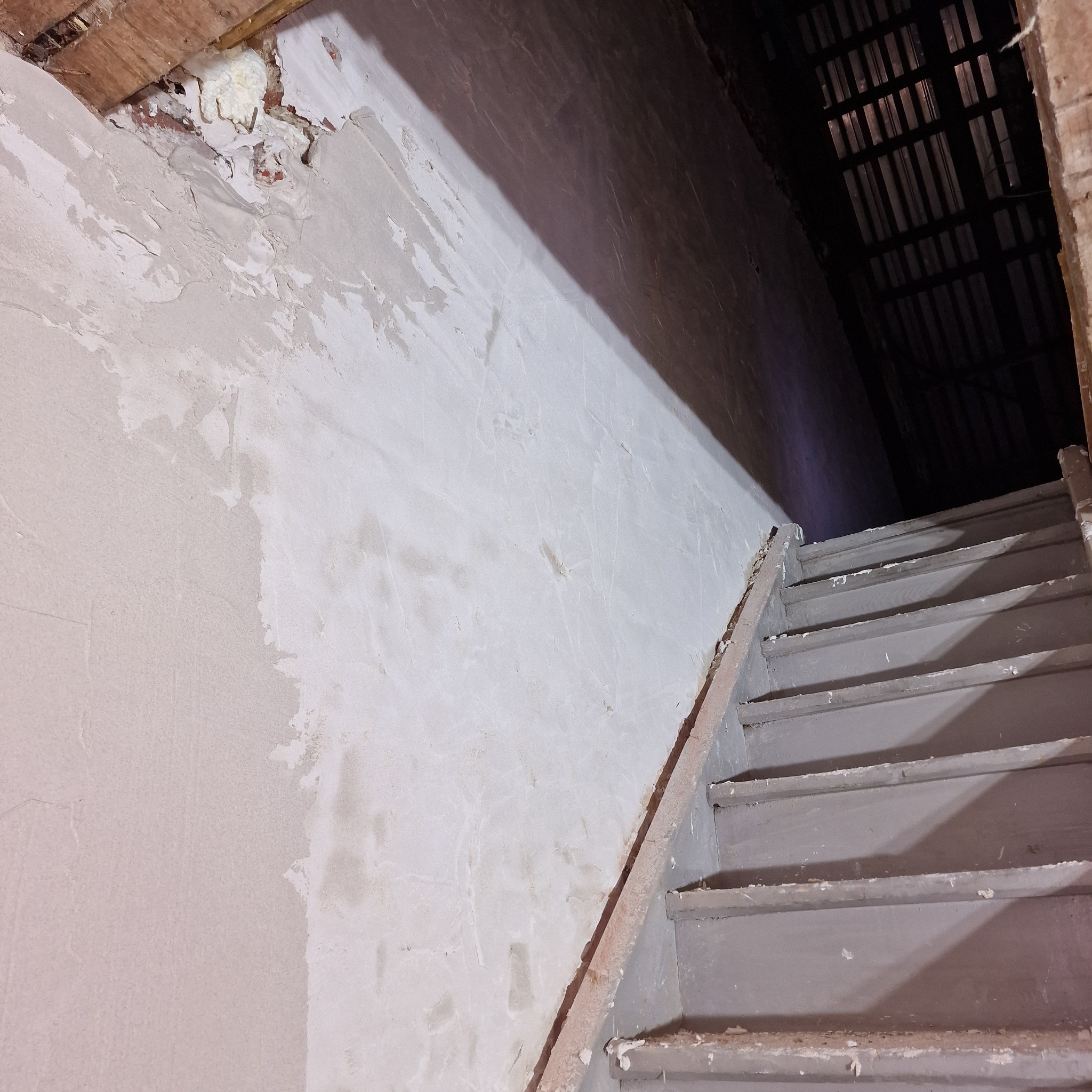 Ściana w trakcie szpachlowania z widocznymi śladami po łataniu ubytków oraz fragmentem drewnianych schodów w ciemnym korytarzu.
