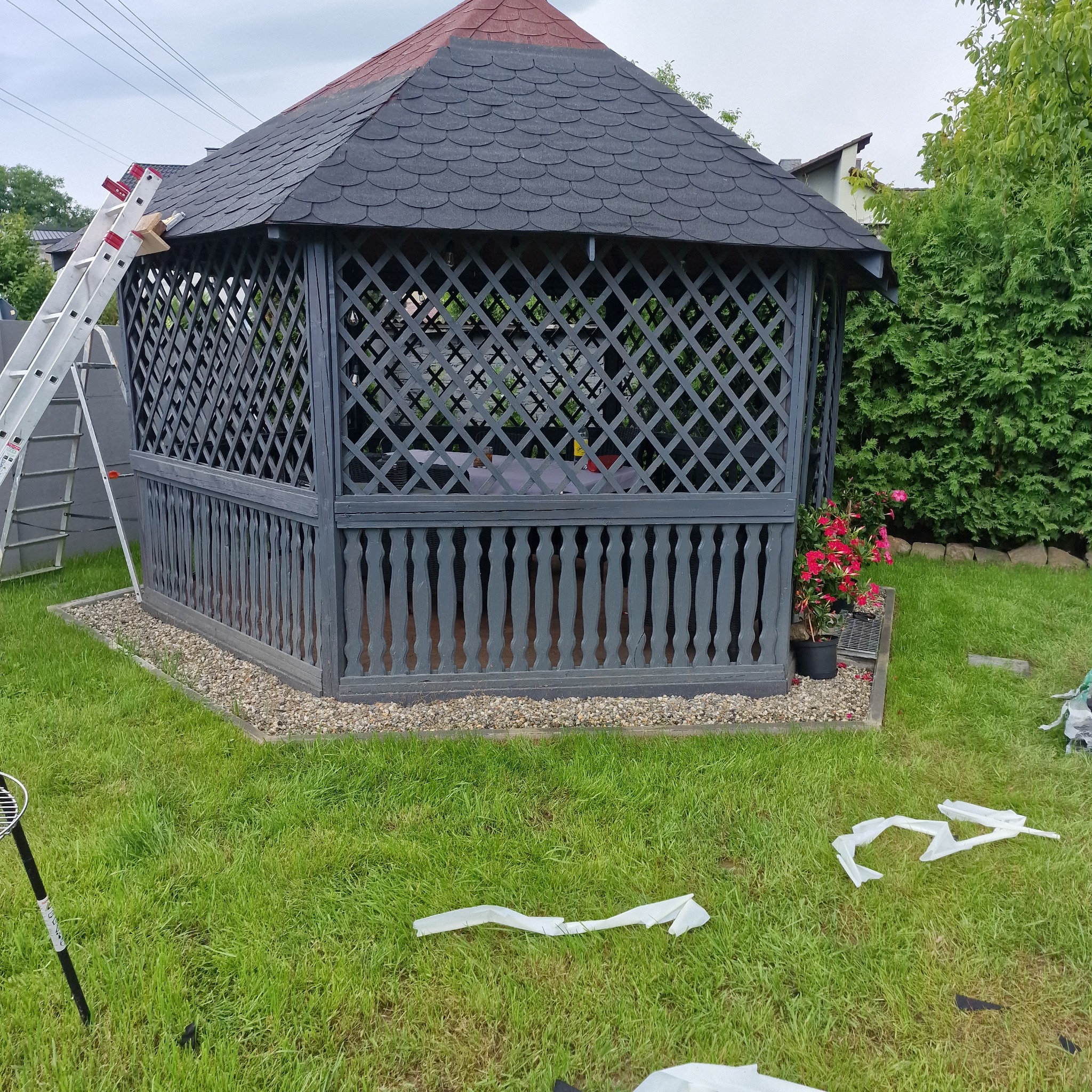 Drewniana altana ogrodowa w trakcie budowy, z rozstawioną drabiną opartą o dach, otoczona trawnikiem i elementami wykończeniowymi.