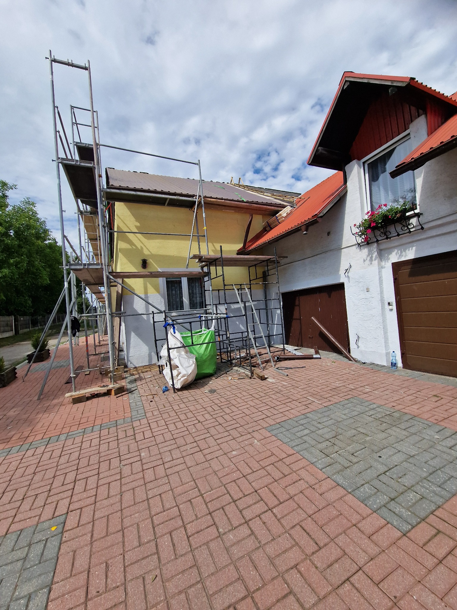 Dom jednorodzinny w trakcie prac remontowych elewacji i dachu, widoczne rusztowania, worki z materiałami budowlanymi i częściowo odnowiona fasada.