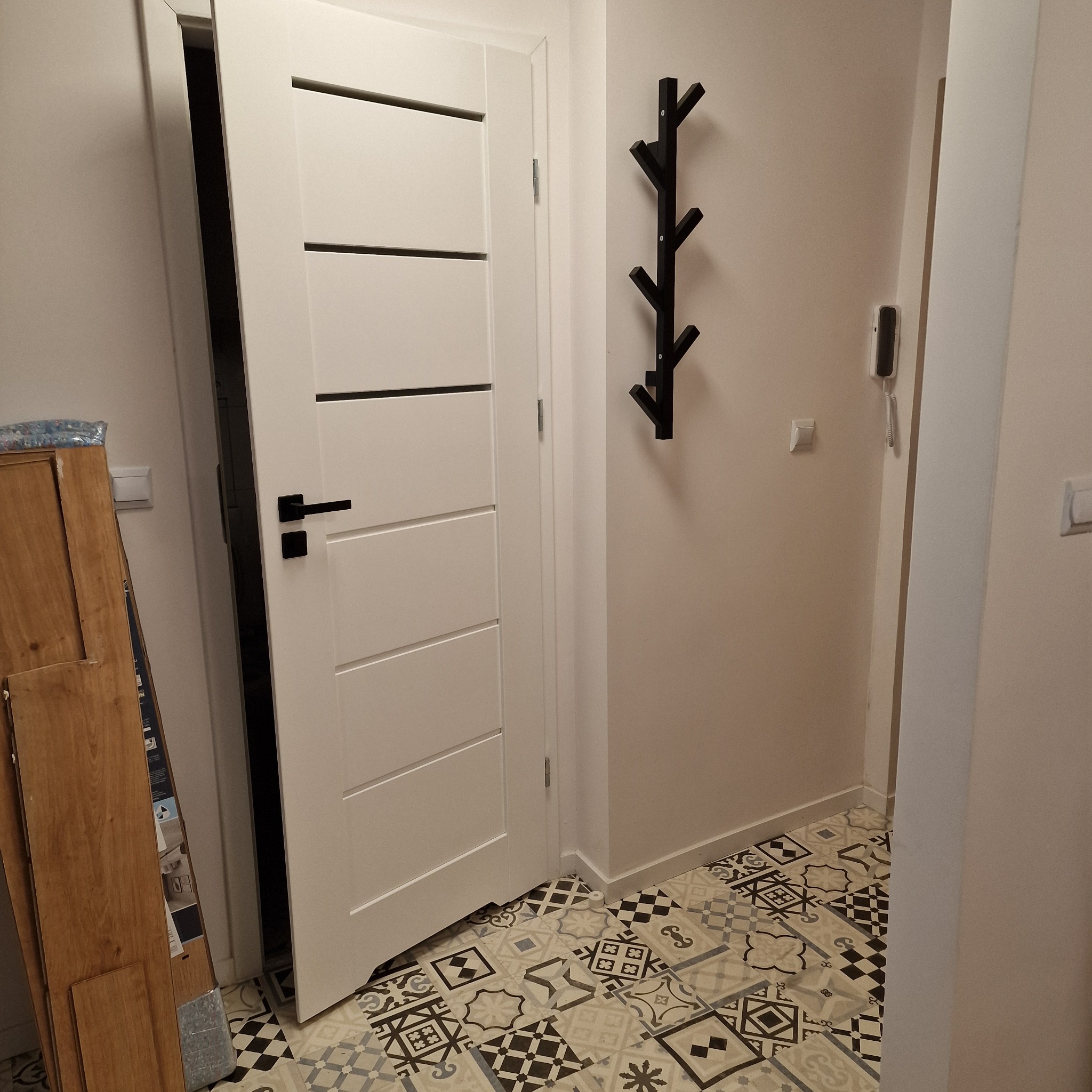 Nowoczesne, białe drzwi wewnętrzne z czarną klamką i poziomymi frezowaniami, zamontowane w przedpokoju z wzorzystą podłogą, obok ściany z wieszakiem na ubrania.