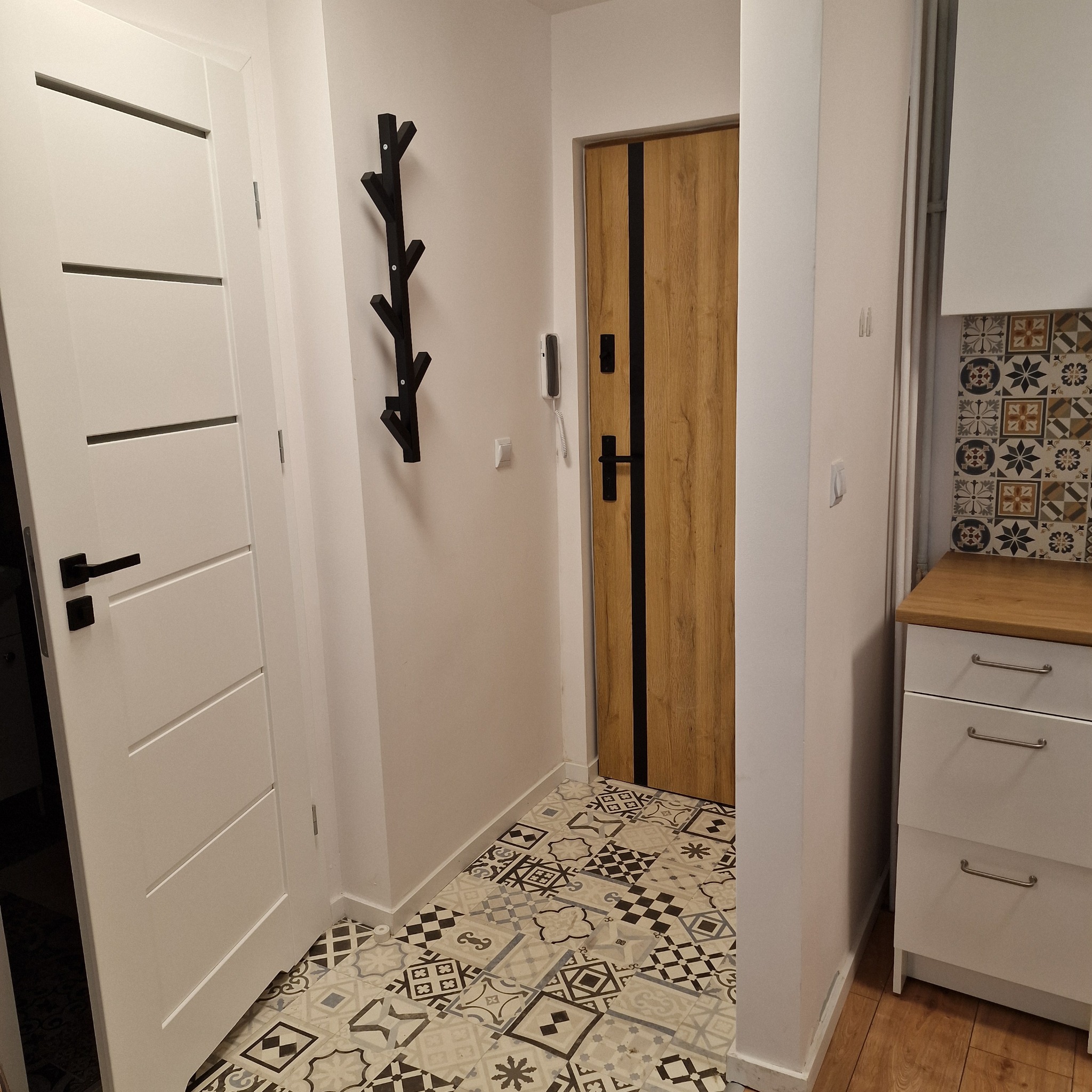 Wykończony przedpokój z drewnopodobnymi drzwiami wejściowymi z czarnymi wstawkami, białe drzwi wewnętrzne z poziomymi frezowaniami, wzorzysta podłoga w stylu patchwork i czarny wieszak na ścianie.
