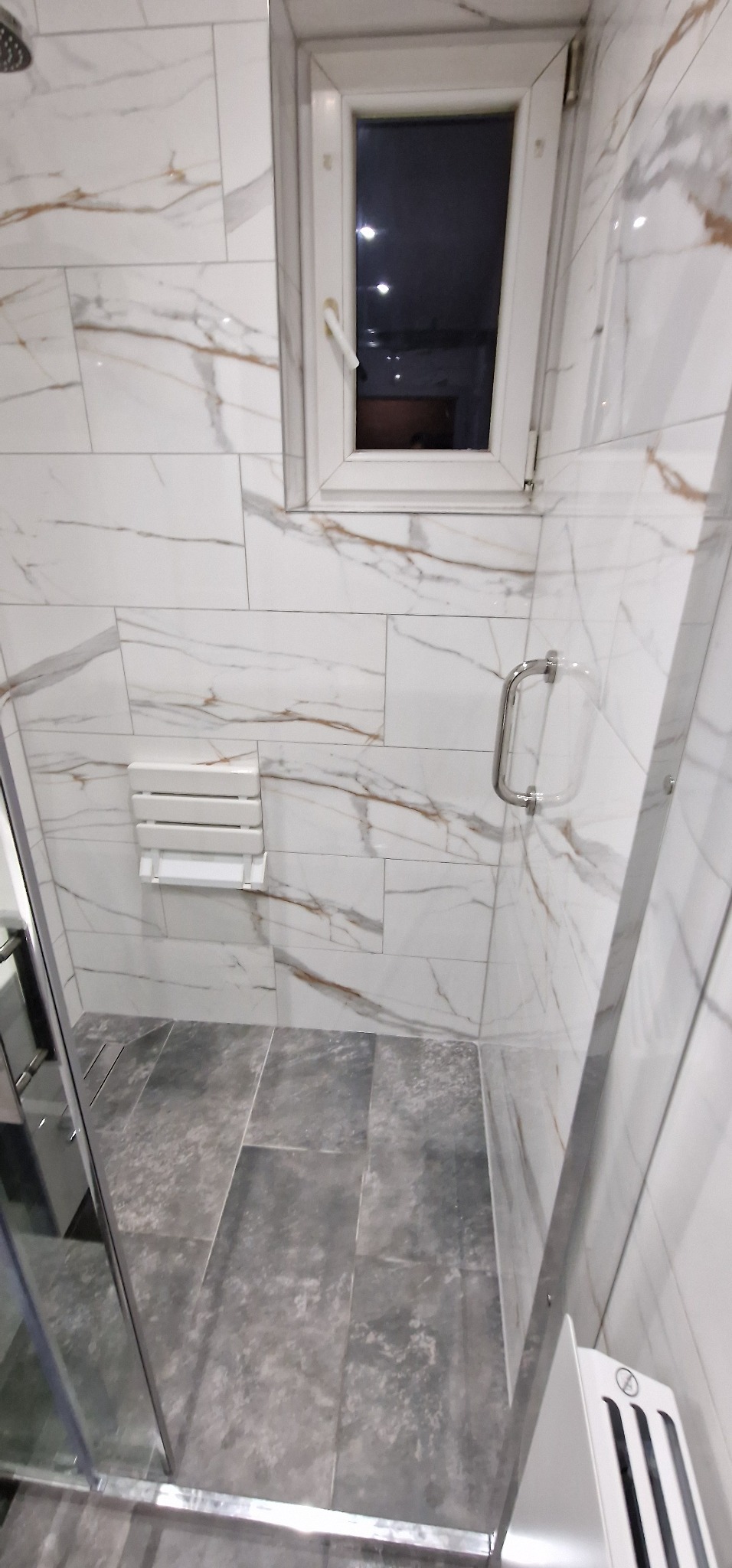 Nowoczesna kabina prysznicowa z białymi płytkami imitującymi marmur i szarymi płytkami podłogowymi, składane siedzisko prysznicowe zamontowane na ścianie.