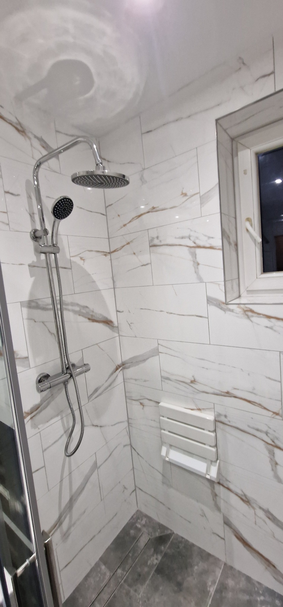 Nowoczesna kabina prysznicowa z deszczownicą i marmurowymi płytkami, składane siedzisko prysznicowe zamontowane na ścianie, okno z białymi ramami.