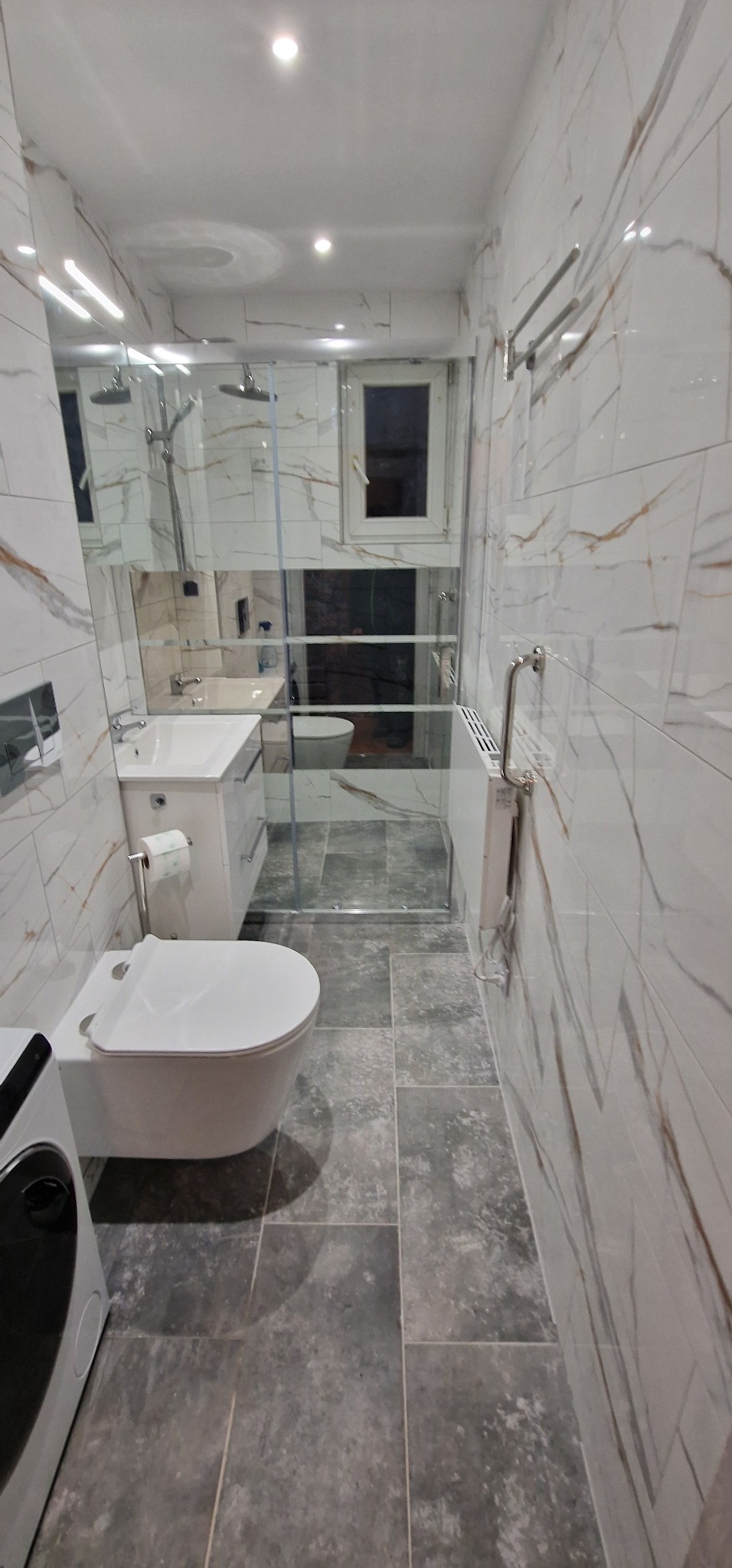 Wąska łazienka z białymi płytkami imitującymi marmur z brązowymi żyłkami, szarą podłogą z płytek, podwieszaną toaletą, umywalką z szafką, grzejnikiem drabinkowym i kabiną prysznicową ze szklaną...