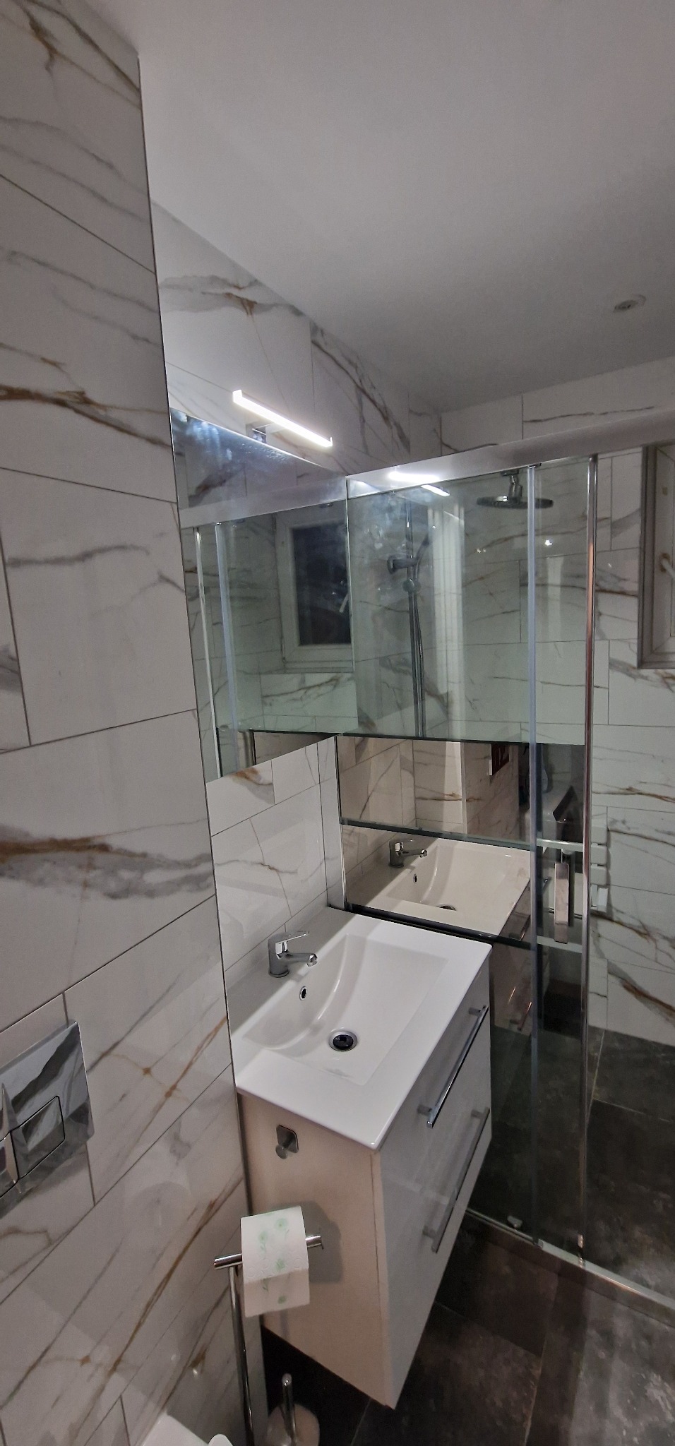Biała umywalka z szafką, chromowana bateria i marmurowe płytki w wyremontowanej łazience z kabiną prysznicową.